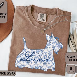 Pu&ograve; includere: T-shirt color espresso con un design floreale blu e bianco di un cane Scottie. Il nome del cane, "Luna", &egrave; scritto in corsivo. La maglietta &egrave; etichettata "Super Soft" e "Comfort Colors".