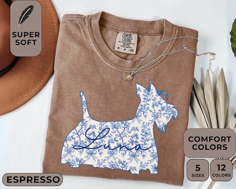 Pu&ograve; includere: T-shirt color espresso con un design floreale blu e bianco di un cane Scottie. Il nome del cane, "Luna", &egrave; scritto in corsivo. La maglietta &egrave; etichettata "Super Soft" e "Comfort Colors".