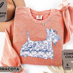 Pu&ograve; includere: Felpa color terracotta con grafica di cane floreale. La silhouette del cane &egrave; riempita con un motivo floreale blu e bianco e il nome "Luna" in corsivo. La maglietta ha il testo "Super Soft" e "Comfort Colors".
