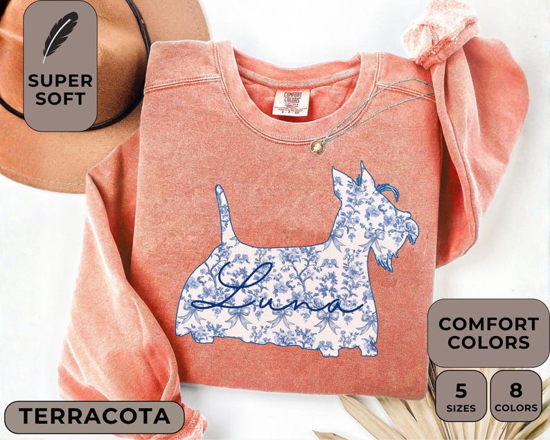 Pu&ograve; includere: Felpa color terracotta con grafica di cane floreale. La silhouette del cane &egrave; riempita con un motivo floreale blu e bianco e il nome "Luna" in corsivo. La maglietta ha il testo "Super Soft" e "Comfort Colors".