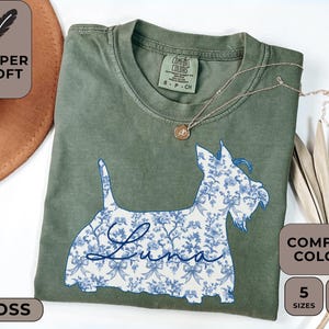 Pu&ograve; includere: T-shirt verde muschio con un disegno floreale di cane blu e bianco. Il nome del cane, "Luna", &egrave; scritto in corsivo. La maglietta ha le parole "Super Soft" e "Comfort Colors" stampate sopra. Disponibile in 5 taglie e 12 colori.