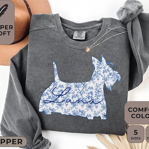 Pu&ograve; includere: Una felpa grigio scuro con un design di Scottish Terrier floreale blu. La parola "Luna" &egrave; scritta in corsivo sul corpo del cane. La felpa ha un girocollo e maniche lunghe. L'immagine include anche il testo "Super Soft" e "Comfort Colors".