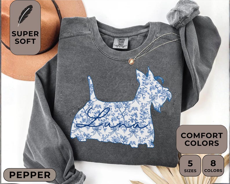 Pu&ograve; includere: Una felpa grigio scuro con un design di Scottish Terrier floreale blu. La parola "Luna" &egrave; scritta in corsivo sul corpo del cane. La felpa ha un girocollo e maniche lunghe. L'immagine include anche il testo "Super Soft" e "Comfort Colors".