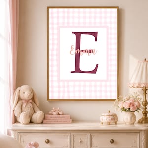 Può includere: Una stampa artistica da parete rettangolare in una cornice dorata con una grande lettera E bordeaux scuro e il nome Emma scritto in un carattere corsivo rosa chiaro al centro, su sfondo bianco con un bordo a quadretti vichy rosa e bianco.