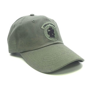 May include: Olive green baseball cap with a curved brim. The cap features a patch with the text "JUNGLE EXPERT" and a graphic. An American flag is embroidered on the side. A casual hat for outdoor activities.