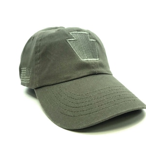 Puede incluir: Gorra de béisbol verde oliva con visera curva. La gorra presenta un parche gris con un diseño de clave de bóveda en la parte delantera y una pequeña bandera estadounidense en el lateral. Fabricada en tejido resistente.