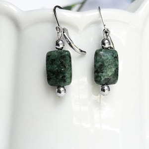 May include: A pair of dangle earrings featuring dark green, rectangular beads with silver-colored accents. The earrings are displayed against a white background with blue floral accents, showcasing the jewelry's design and color.