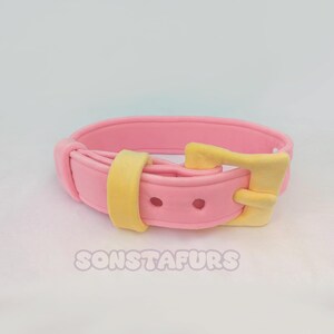 May include: A pink collar with a yellow buckle and loop. The collar has two small holes and the text "SONSTAFURS" is visible at the bottom. The collar is likely for a pet.