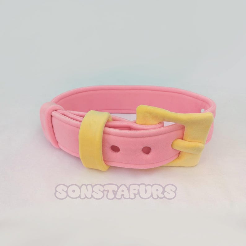 May include: A pink collar with a yellow buckle and loop. The collar has two small holes and the text "SONSTAFURS" is visible at the bottom. The collar is likely for a pet.
