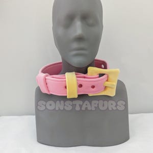 May include: A pink collar with a yellow buckle is displayed on a gray mannequin head. The collar has a rounded shape with a yellow strap and buckle. The text "SONSTAFURS" is visible at the base of the mannequin.