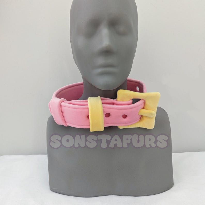 May include: A pink collar with a yellow buckle is displayed on a gray mannequin head. The collar has a rounded shape with a yellow strap and buckle. The text "SONSTAFURS" is visible at the base of the mannequin.