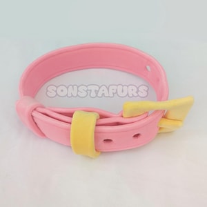 May include: A pink collar with a yellow buckle and accents. The collar has the text "SONSTAFURS" printed on it. The collar is made of a soft material.