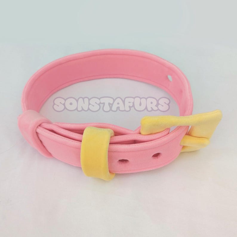 May include: A pink collar with a yellow buckle and accents. The collar has the text "SONSTAFURS" printed on it. The collar is made of a soft material.