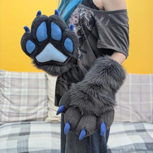 May include: Gray faux fur paw gloves with blue paw pads and claws. The gloves are designed to resemble animal paws, with a plush, textured appearance. The gloves are a unique accessory for cosplay or costume events.