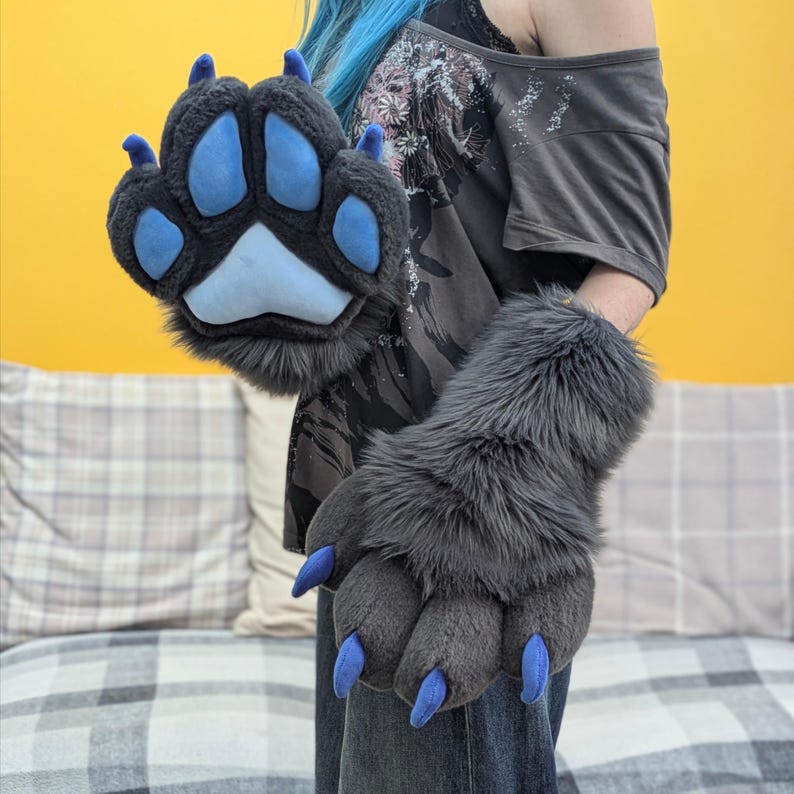 May include: Gray faux fur paw gloves with blue paw pads and claws. The gloves are designed to resemble animal paws, with a plush, textured appearance. The gloves are a unique accessory for cosplay or costume events.