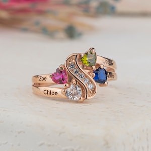 Puede incluir: Un anillo de oro con un diseño de onda y cinco piedras preciosas. El anillo está grabado con los nombres "Chloe", "Zoe" y "Jayde".