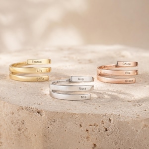 Puede incluir: Tres anillos envolventes en tonos oro, plata y oro rosa. Cada anillo presenta tres bandas apiladas, con los nombres Emma, Lily y Sofia en el anillo dorado; Ava, Nora y Mia en el anillo plateado; y Chloe, Ella y Grace en el anillo de oro rosa.