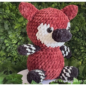 May include: A handmade crochet fox plushie in shades of red, white, and black. The fox has a textured body, white face markings, and black and white checkered paws and feet. The plushie is set against a backdrop of green foliage.