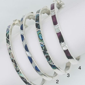 Può includere: Quattro braccialetti d'argento con diversi disegni di intarsi in madreperla di abalone. I braccialetti sono impilati uno sopra l'altro, con il braccialetto superiore con un intarsio viola, il secondo braccialetto con un intarsio blu, il terzo braccialetto con un intarsio verde e il braccialetto inferiore con un intarsio multicolore. I braccialetti sono numerati da 1 a 4.