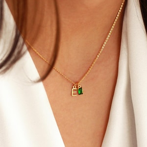 May include: A gold-colored necklace with a delicate chain. The pendant features the letter "E" and a small green rectangular gemstone. The necklace is worn against a white fabric background.