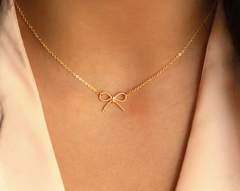 May include: A delicate gold-colored necklace featuring a small bow pendant. The bow has a textured design and is suspended from a fine chain. The necklace is shown against a light pink background.