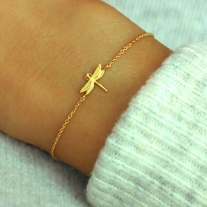May include: A gold-coloured bracelet with a dragonfly charm. The bracelet has a fine chain and a small, detailed dragonfly pendant. The jewellery is worn on a wrist, with a light-coloured sweater in the background. The bracelet is a simple, elegant accessory.