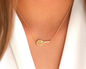May include: A gold-coloured tennis racket pendant necklace. The pendant is a detailed miniature of a tennis racket. The necklace is worn against a white top. The chain is delicate and gold-coloured.