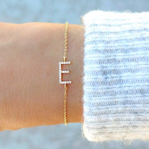 May include: A gold-colored bracelet featuring the letter "E" encrusted with small, clear gemstones. The bracelet is attached to a delicate chain. The background is a soft, blurred gray and white.