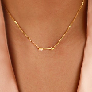 May include: A delicate gold-colored necklace featuring a small arrow pendant. The arrow is horizontally oriented, pointing to the right. The necklace is displayed against a soft, peach-colored background, highlighting the jewelry's simple elegance.