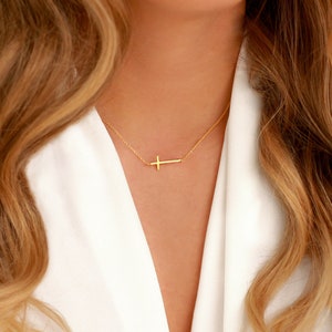 Könnte beinhalten: Eine goldfarbene Halskette mit einem seitlichen Kreuzanhänger. Die Kette und das Kreuz sind vor einem weißen Hintergrund sichtbar. Die Halskette ist ein schlichtes, elegantes Schmuckstück.