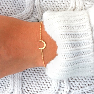 May include: A delicate gold-colored bracelet featuring a crescent moon charm. The bracelet is worn on a wrist, set against a backdrop of a white knitted sweater. The moon charm is a simple, elegant design.