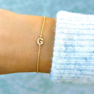 Può includere: Un delicato braccialetto dorato con un design a catena. Il braccialetto presenta un singolo ciondolo con la lettera "G". Il braccialetto è indossato su un polso, con un maglione azzurro e bianco sullo sfondo.