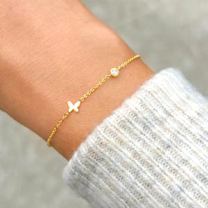 May include: A delicate gold bracelet featuring a small butterfly charm and a single, round, clear gemstone. The bracelet is worn on a wrist, and the gold chain is visible against the skin and a light gray sweater.
