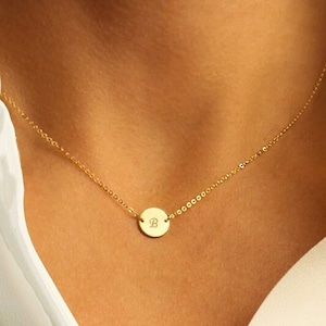 Può includere: Una collana dorata con una catena sottile e un ciondolo circolare. Il ciondolo presenta la lettera "B" in un carattere elegante. La collana è indossata su una camicetta bianca.