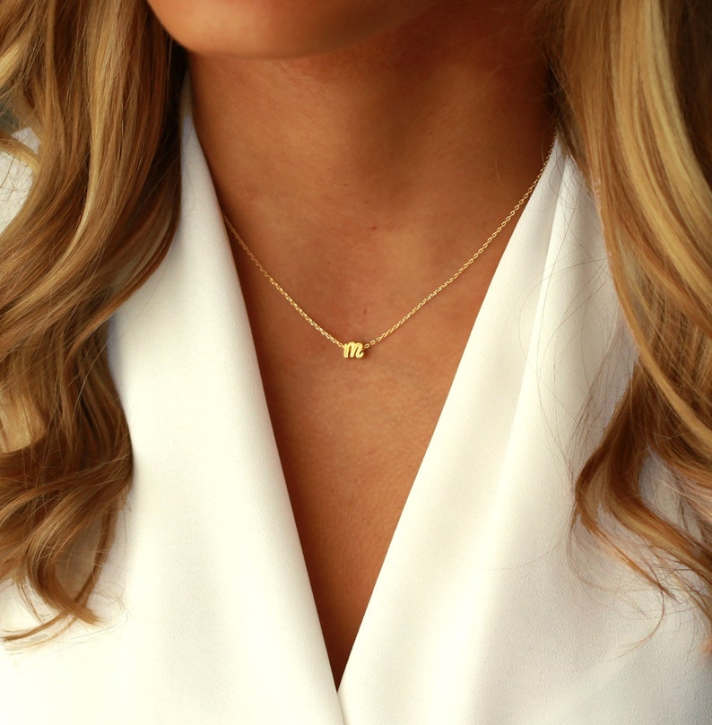 Puede incluir: Un delicado collar de oro con un colgante con la letra "m". El colgante es peque&ntilde;o y est&aacute; centrado en una fina cadena. El collar se lleva sobre una blusa blanca, con cabello rubio enmarcando el cuello.