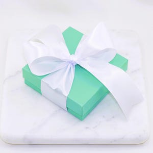 Puede incluir: Una peque&ntilde;a caja de regalo cuadrada, verde claro, atada con un gran lazo de sat&eacute;n blanco. La caja est&aacute; sobre una superficie de m&aacute;rmol blanco, creando una presentaci&oacute;n limpia y elegante. Las cintas del lazo caen sobre la caja.