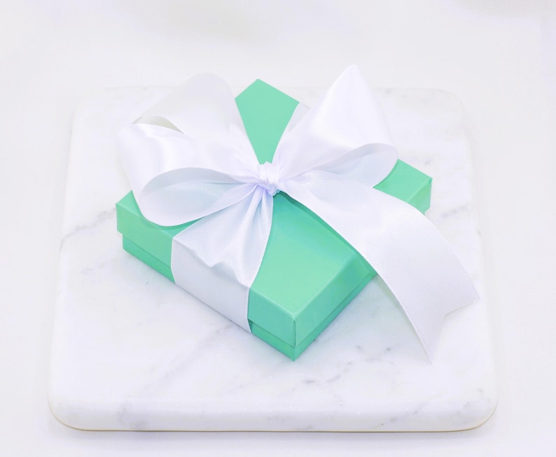 Puede incluir: Una peque&ntilde;a caja de regalo cuadrada, verde claro, atada con un gran lazo de sat&eacute;n blanco. La caja est&aacute; sobre una superficie de m&aacute;rmol blanco, creando una presentaci&oacute;n limpia y elegante. Las cintas del lazo caen sobre la caja.