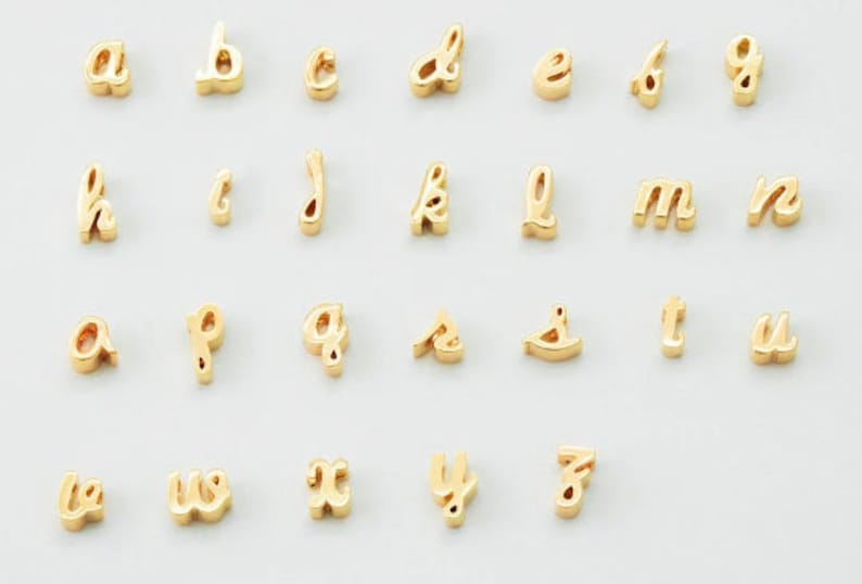 Puede incluir: Una colecci&oacute;n de abalorios dorados con letras min&uacute;sculas del alfabeto, cada uno con una fuente cursiva. Las letras est&aacute;n dispuestas sobre una superficie blanca. Estos abalorios son adecuados para la fabricaci&oacute;n de joyas o manualidades personalizadas.