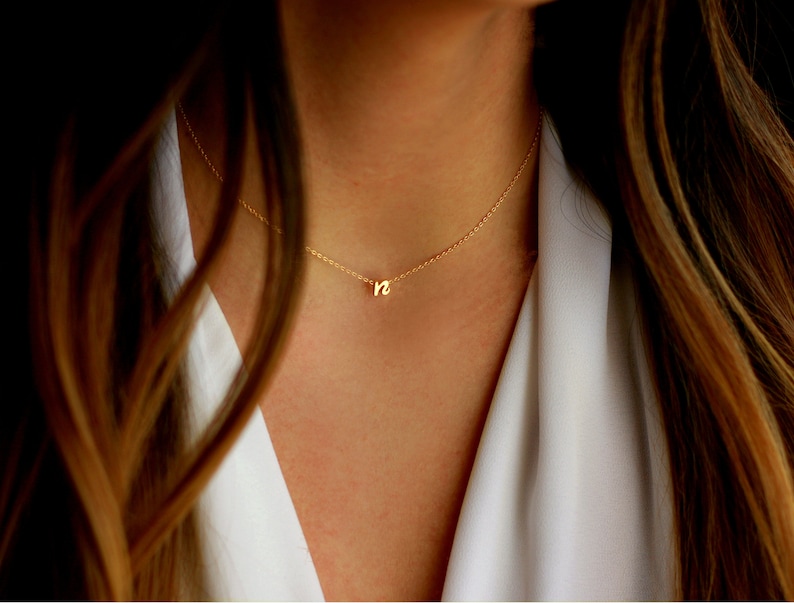 Puede incluir: Un delicado collar dorado con un peque&ntilde;o colgante con la letra "n" en min&uacute;scula. El collar se lleva alrededor del cuello, con la cadena visible sobre una blusa blanca y cabello casta&ntilde;o.