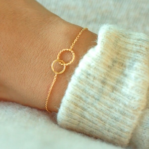 May include: A delicate gold-colored bracelet featuring two interlocking circles. One circle has a textured, beaded design, while the other is smooth. The bracelet is attached to a fine chain, creating a minimalist and elegant jewelry piece.