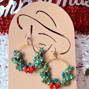 May include: Gold hoop earrings featuring a Christmas wreath design. Each earring has a green wreath with red bow and berries. The earrings are set against a festive background with the word "Christmas".