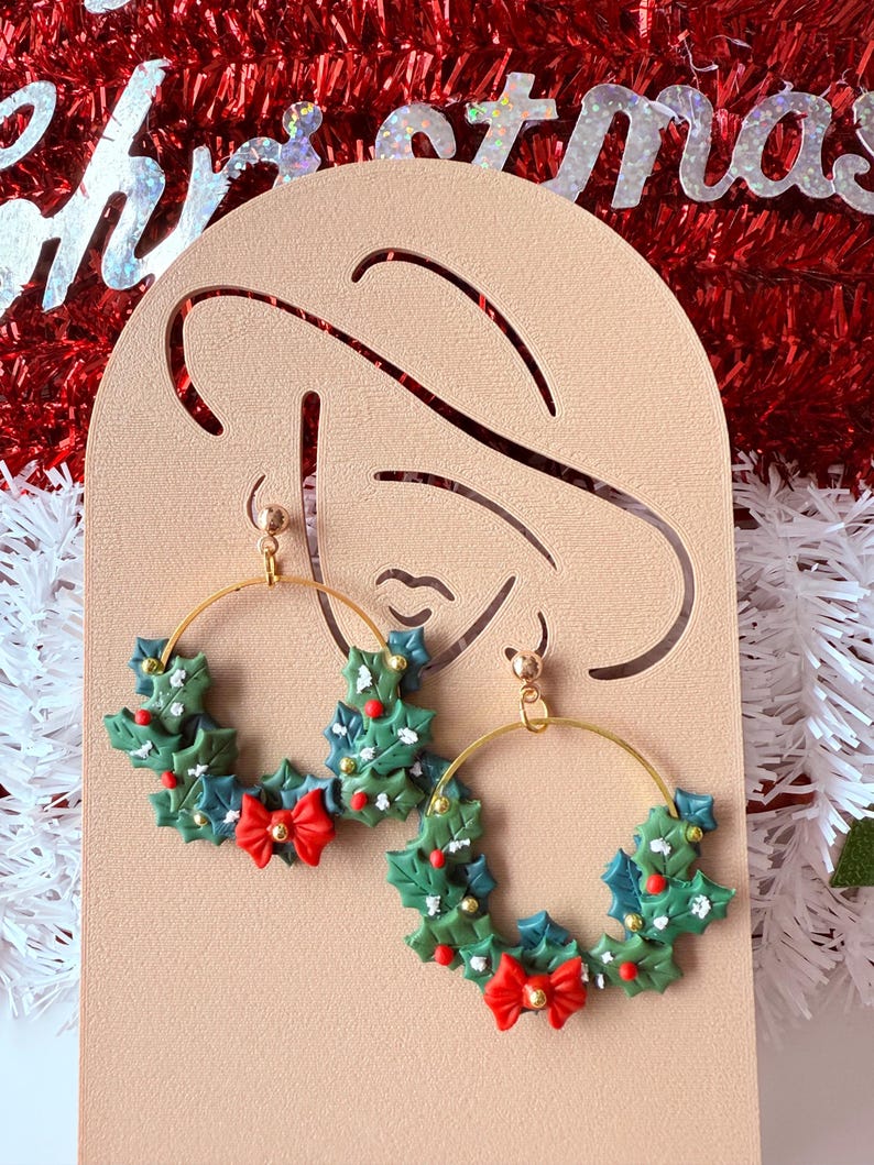 May include: Gold hoop earrings featuring a Christmas wreath design. Each earring has a green wreath with red bow and berries. The earrings are set against a festive background with the word "Christmas".