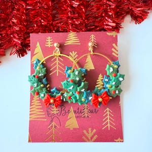 May include: Gold hoop earrings with a festive Christmas holly design. The earrings feature green holly leaves, red berries, and red bow accents. The earrings are displayed on a red card with gold tree patterns and the text "Beautifur Collect-our".