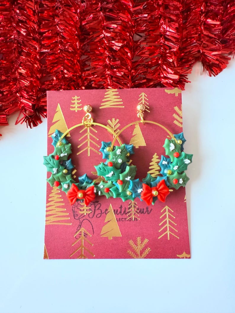 May include: Gold hoop earrings with a festive Christmas holly design. The earrings feature green holly leaves, red berries, and red bow accents. The earrings are displayed on a red card with gold tree patterns and the text "Beautifur Collect-our".