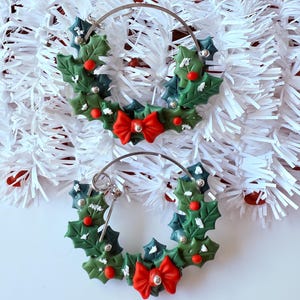May include: A pair of festive hoop earrings. Each earring features a green holly wreath design with red berries, silver accents, and a red bow. The earrings are set against a white background with red and white decorative elements.