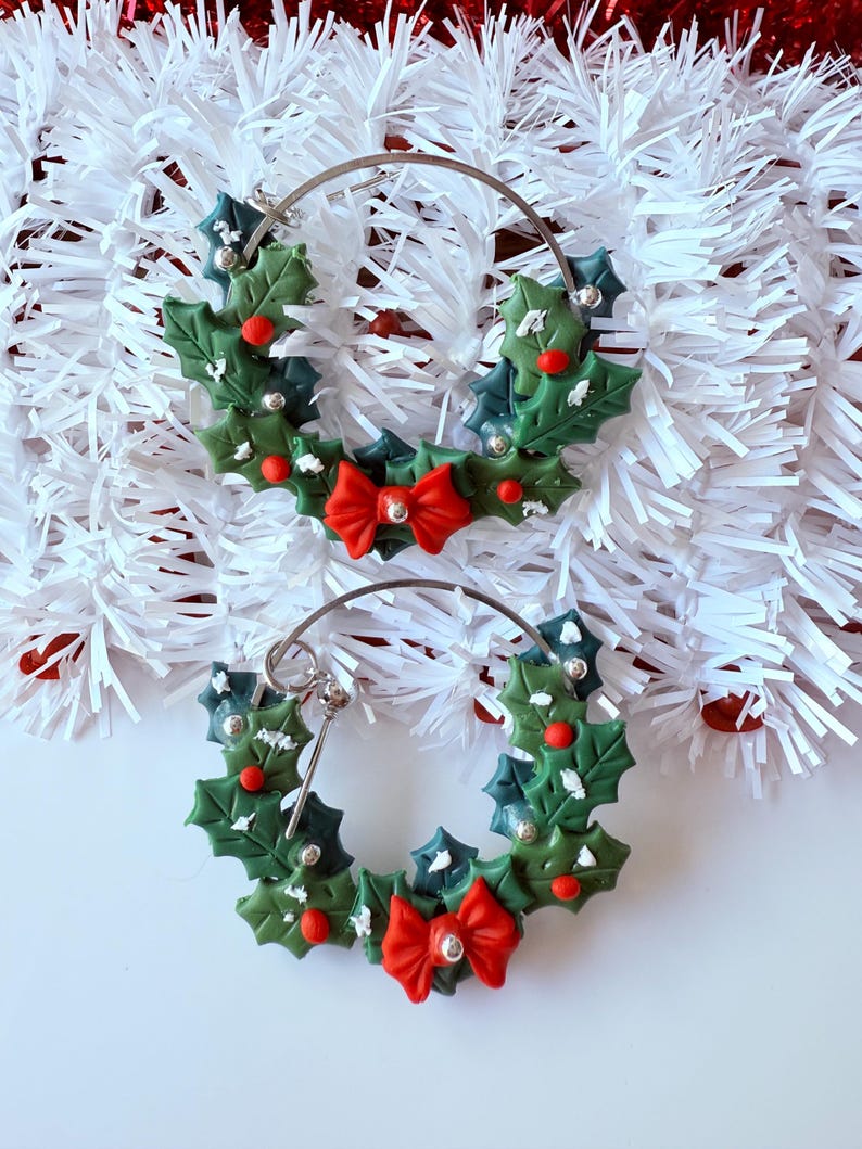 May include: A pair of festive hoop earrings. Each earring features a green holly wreath design with red berries, silver accents, and a red bow. The earrings are set against a white background with red and white decorative elements.