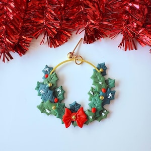 May include: A festive Christmas wreath earring with a gold hoop. The wreath is made of green and blue leaves, red berries, and a red bow. The earring is a unique holiday accessory.