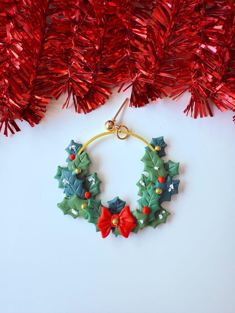 May include: A festive Christmas wreath earring with a gold hoop. The wreath is made of green and blue leaves, red berries, and a red bow. The earring is a unique holiday accessory.