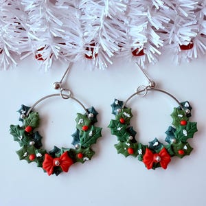 May include: A pair of hoop earrings with a Christmas wreath design. The earrings feature green holly leaves, red berries, and a red bow. The hoops are silver-colored and have a post closure. The background is white with white garland.