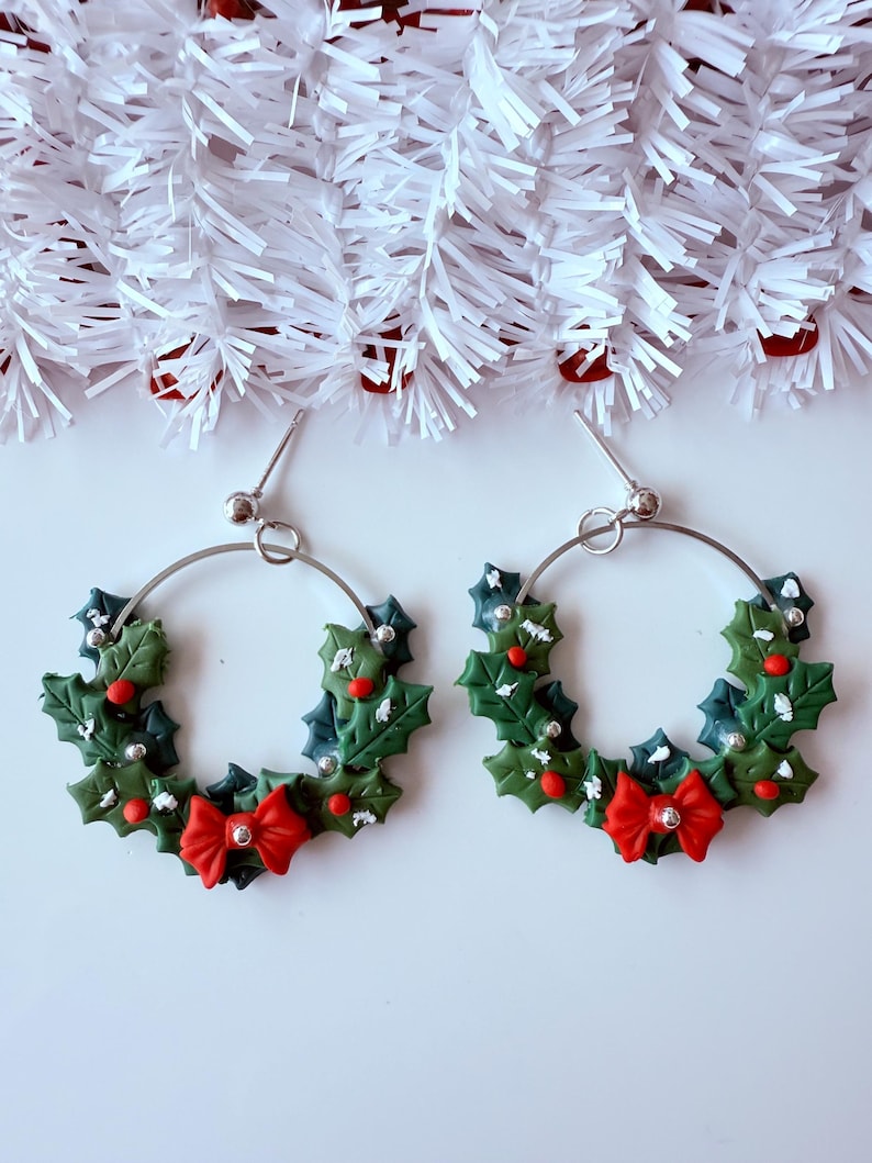 May include: A pair of hoop earrings with a Christmas wreath design. The earrings feature green holly leaves, red berries, and a red bow. The hoops are silver-colored and have a post closure. The background is white with white garland.